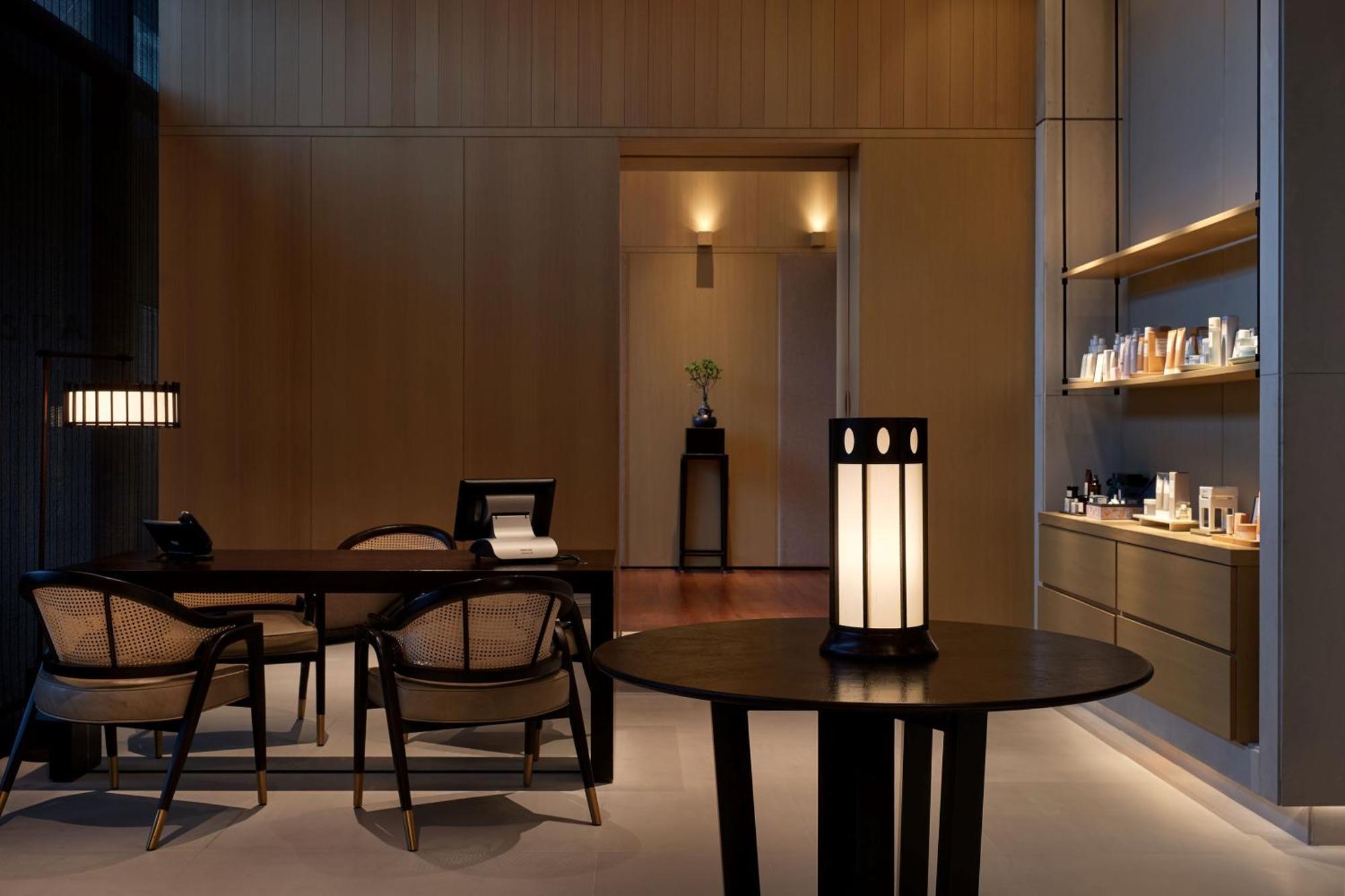 더 루마 호텔 앤 레지던스 쿠알라룸푸르 외부 사진 The photo shows a modern, minimalist interior space. It features a wooden wall with soft lighting, enhancing a calm atmosphere. In the foreground, there is a round table with a uniquely designed lamp on it. Surrounding the table, there are stylish wo